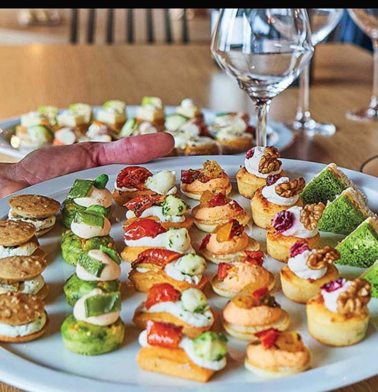 A wide, close-up shot of a large, circular white plate filled with an assortment of savory appetizers, also known as canapés. The appetizers are arranged in neat rows. One row consists of small round bread with what looks like a white cheese spread and two small pieces of tomato. Another row has a green circular base topped with a white spread and a garnish. A third row has tiny puff pastries topped with different spreads, including one with a walnut on top. In the background, there is a second plate of appetizers, along with wine glasses and a hand holding the first plate.