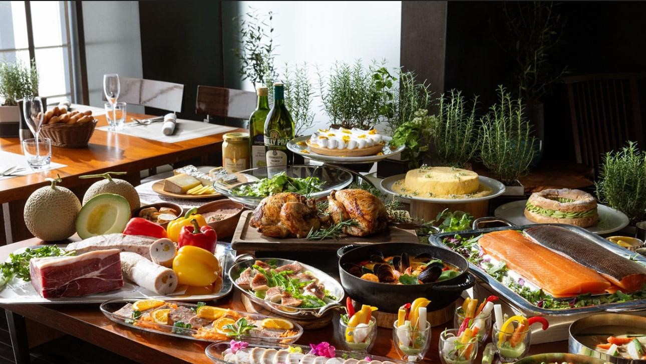 A long buffet table is laden with a lavish spread of food, including whole roasted chickens, a large salmon fillet, a paella dish, and various salads and desserts. The table is also decorated with fresh herbs, bread, bottles of wine, and other drinks. In the background, a dining table is set with glasses and silverware, with natural light coming from a window.