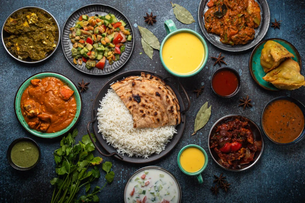 A top-down shot of a variety of Indian dishes arranged on a dark, textured background. The spread includes a large plate of rice and flatbread in the center, surrounded by bowls of different curries, a fresh salad, samosas, sauces, and scattered spices like star anise.