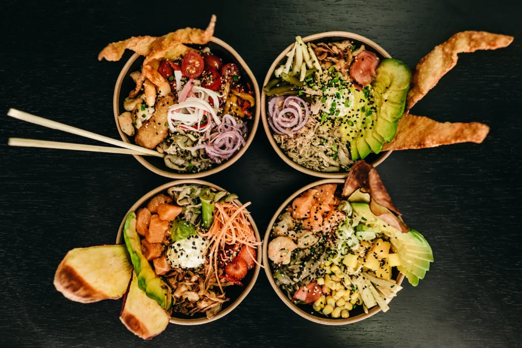 A top-down shot of four paper bowls filled with different ingredients, arranged in a square on a dark, textured surface. Each bowl is a poke bowl variation, filled with rice, protein, and a vibrant mix of vegetables, fruits, and sauces. Ingredients visible include salmon, shrimp, avocado, corn, cherry tomatoes, and various garnishes like seeds and fresh herbs. One bowl has a pair of wooden chopsticks resting on it, and several bowls have a garnish of what appears to be a fried wonton.