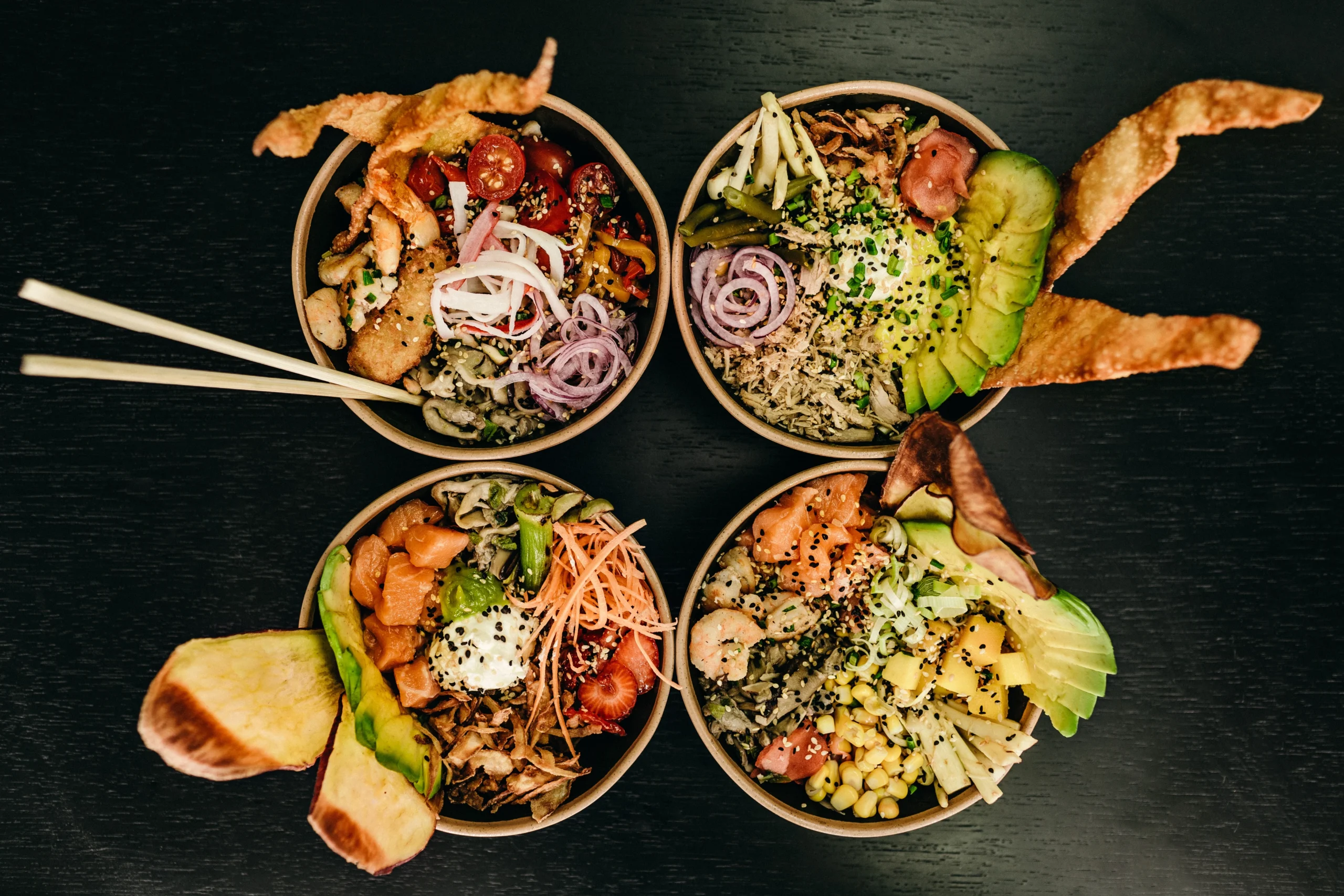 A top-down shot of four paper bowls filled with different ingredients, arranged in a square on a dark, textured surface. Each bowl is a poke bowl variation, filled with rice, protein, and a vibrant mix of vegetables, fruits, and sauces. Ingredients visible include salmon, shrimp, avocado, corn, cherry tomatoes, and various garnishes like seeds and fresh herbs. One bowl has a pair of wooden chopsticks resting on it, and several bowls have a garnish of what appears to be a fried wonton.