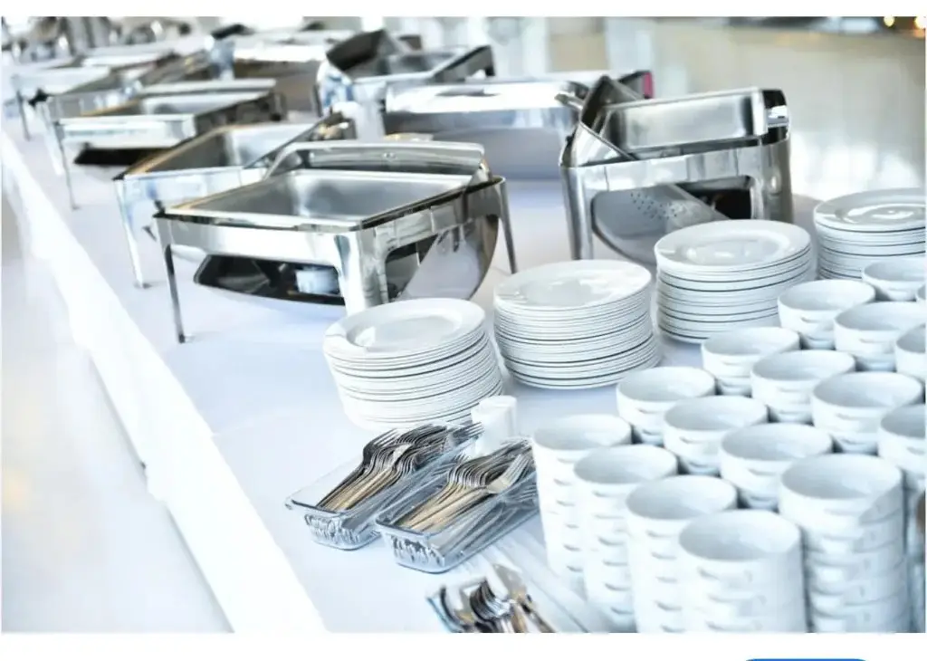 A wide-angle shot of a clean buffet table setup for an event. In the foreground, neat stacks of white ceramic plates and bowls are arranged next to trays of forks and spoons. In the background, a long line of empty, shiny metal chafing dishes is set up on the white-clothed table, ready for food.