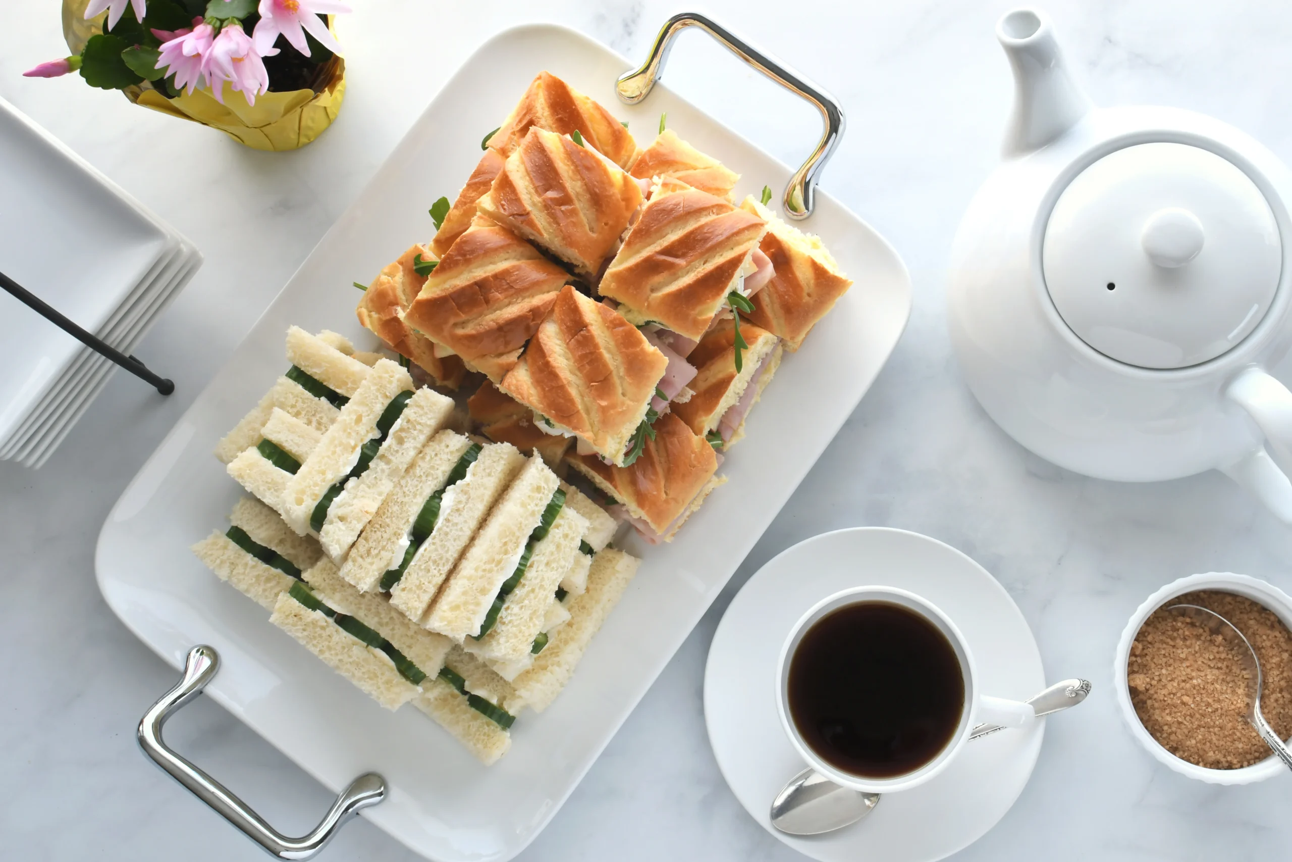 A high-angle shot of a platter of sandwiches on a marble table, set for afternoon tea. The platter has two types of sandwiches: a stack of small, crustless finger sandwiches filled with a green herb spread, and another stack of triangular sandwiches on toasted bread. The platter is on a white ceramic tray with metal handles. A white teapot, a cup of coffee, and a bowl of brown sugar with a spoon are also on the table. A potted pink flowering plant is in the upper left corner.