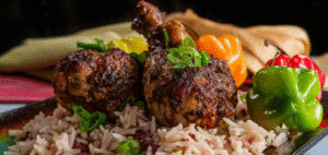A close-up shot of two juicy, roasted chicken legs, also known as drumsticks, covered in a dark, savory sauce. They are resting on a bed of white rice mixed with red beans. The dish is garnished with sliced green onions and accompanied by colorful bell peppers in the background.