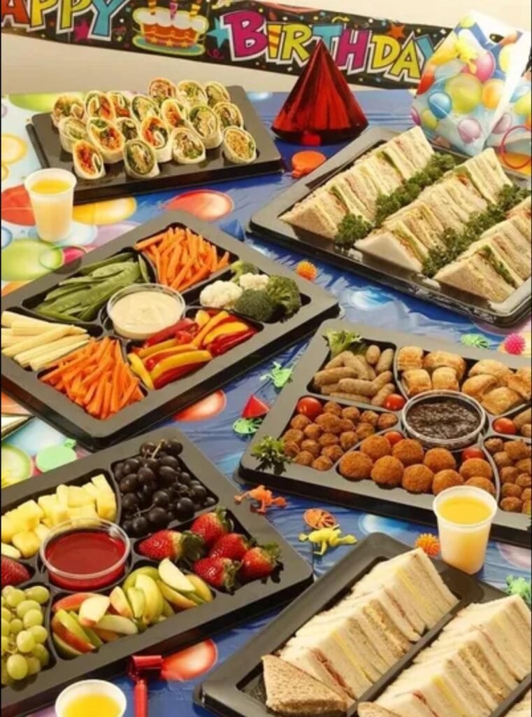 A high-angle shot of a table covered with a birthday-themed tablecloth and a "Happy Birthday" banner. Several black plastic platters are filled with various party foods. Two platters contain cut sandwiches, two have a variety of finger foods like meatballs and sausage rolls, one has a vegetable assortment with dip, and one has fruit. A platter of wraps cut into slices is also visible. Small cups of juice are placed on the table, along with a party hat and a small bag of balloons.