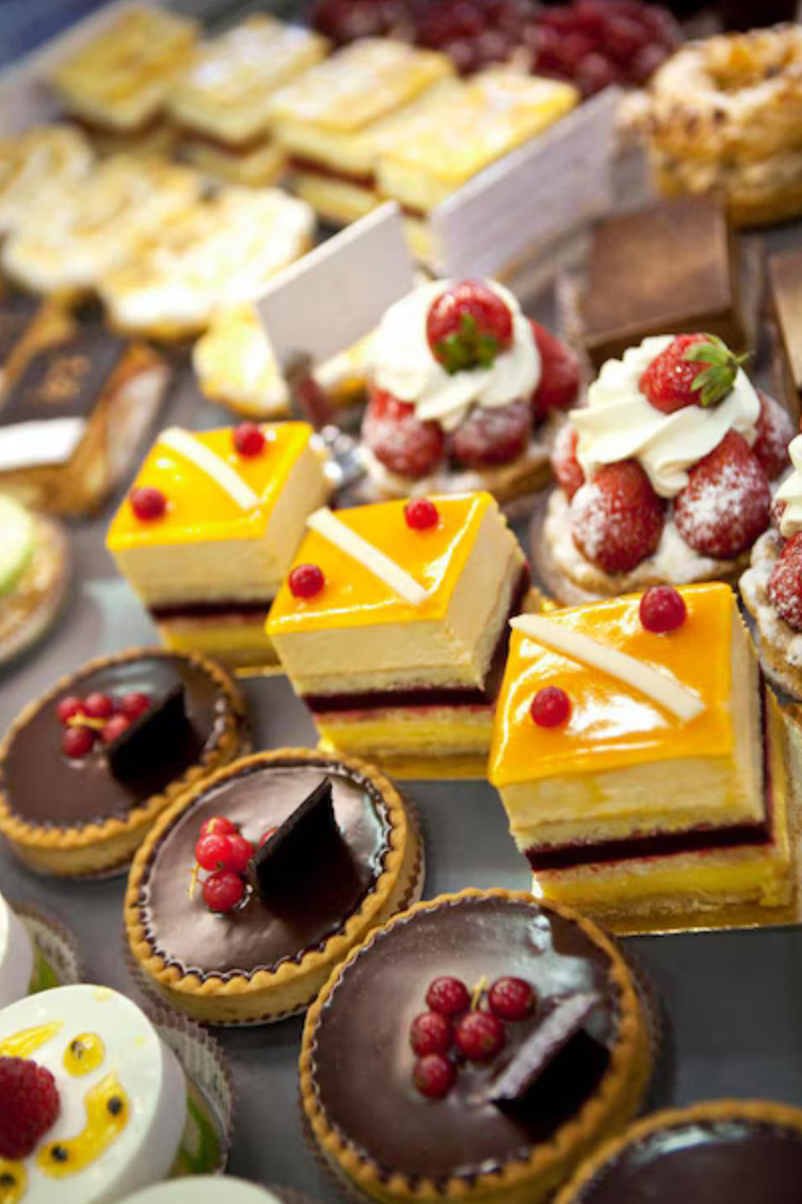 A close-up, high-angle shot of a display case filled with a variety of small, colorful desserts. The photo is taken from above, with rows of pastries and tarts visible. In the foreground, there are three chocolate tarts topped with red currants and a small piece of dark chocolate, and three square layered cakes with a bright yellow glaze and red fruit on top. In the background, there are pastries topped with whipped cream and strawberries.