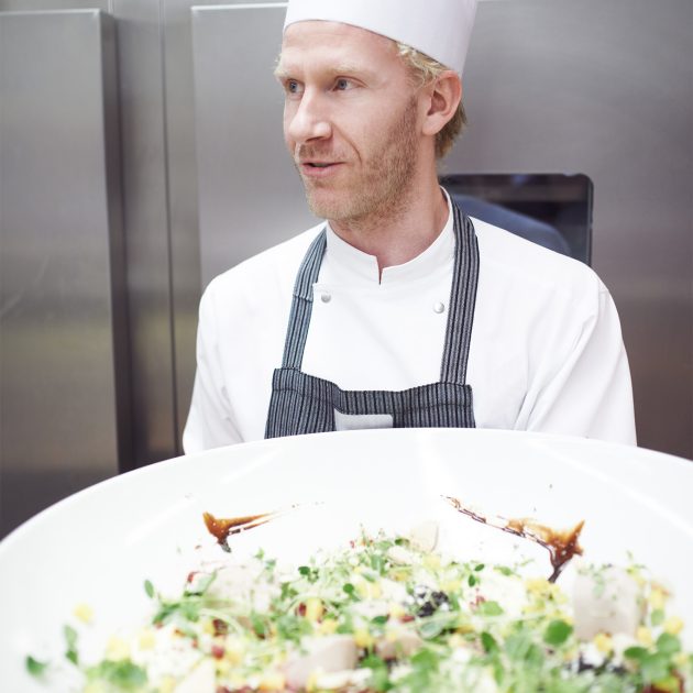 chef1 A chef with blonde hair and a beard, wearing a white chef's hat and coat with a striped apron, stands in a professional kitchen. He is holding a large white platter of a beautifully plated salad with greens, various toppings, and a dark sauce drizzled on top. He is looking off to the side, away from the camera. The background shows stainless steel kitchen equipment.
